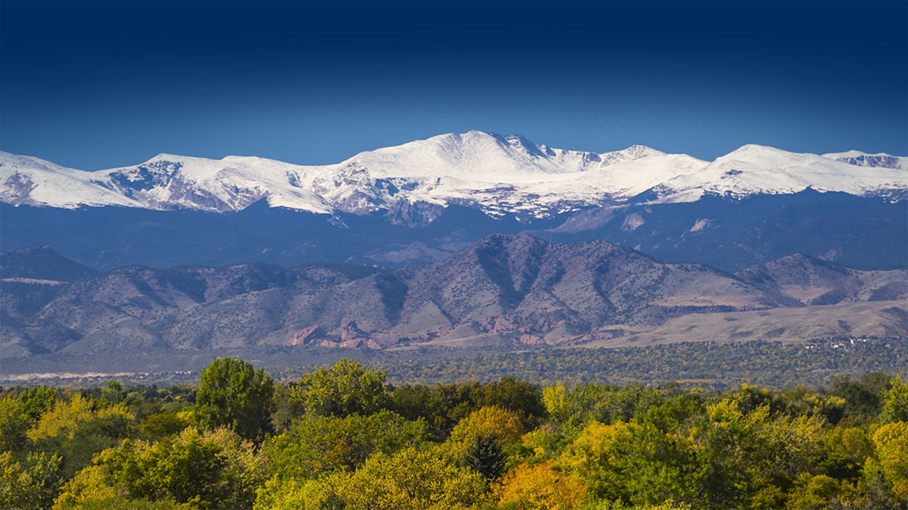 A view of the Rocky Mountains with snow-capped mountains, green and brown foothills and bright green trees in the valley.