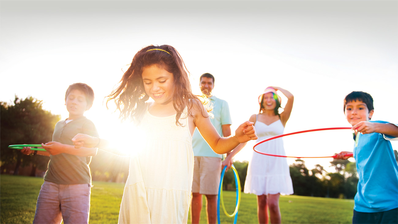 A girl with long brown hair and yellow dress stands in the sunlight in front of her family holding hula hoops in a field.